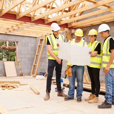 Builder On Building Site Looking At Plans With Apprentices