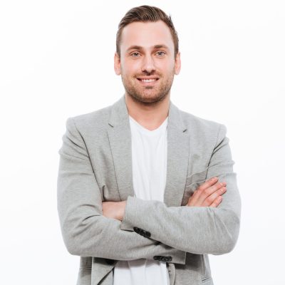 Portrait of successful man having stubble posing on camera with broad smile keeping arms folded isolated over white background